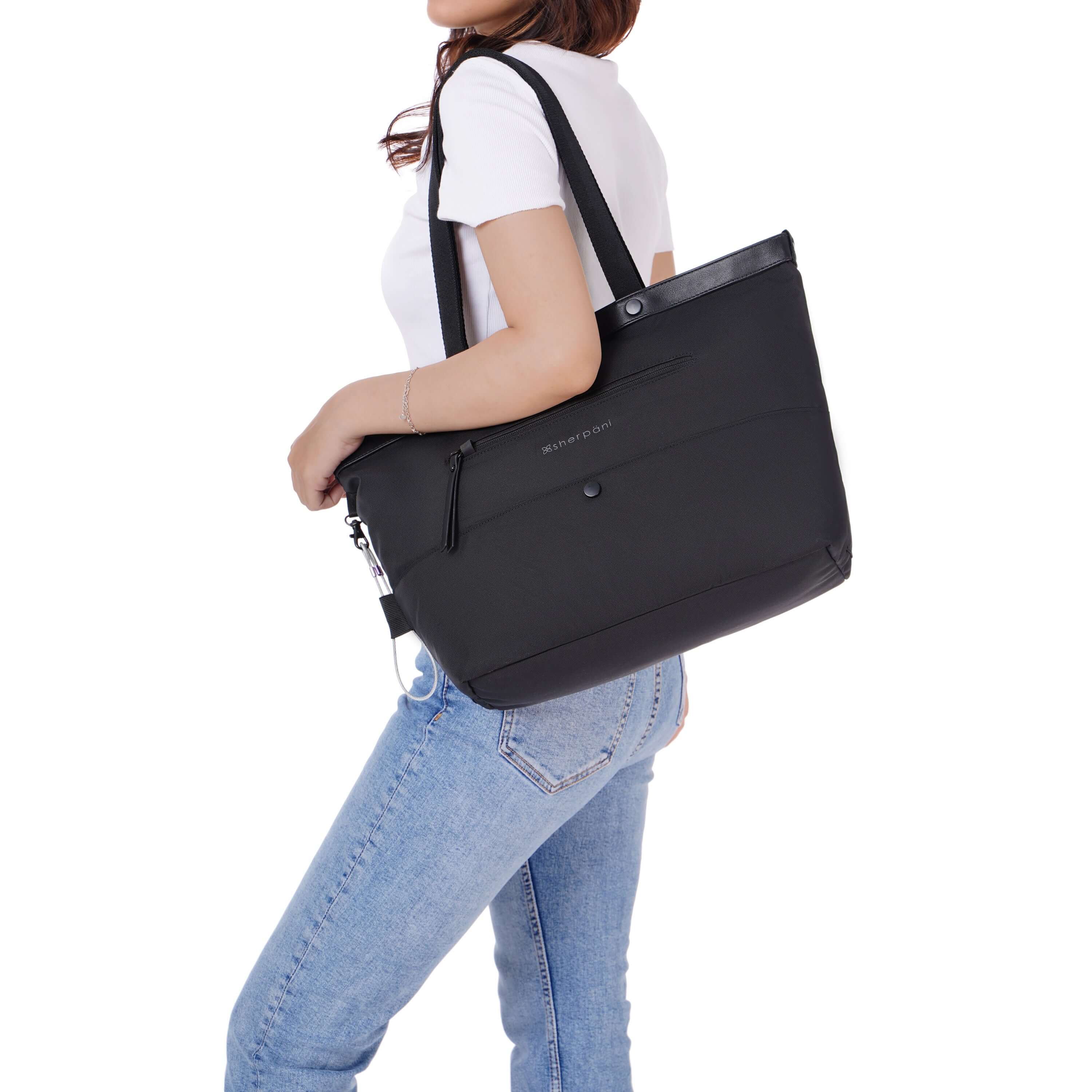 A brown haired model is facing the side. She is wearing a white tee shirt and jeans. Sherpani&#39;s Anti-Theft tote the Cali AT in Carbon sits on her shoulder.
