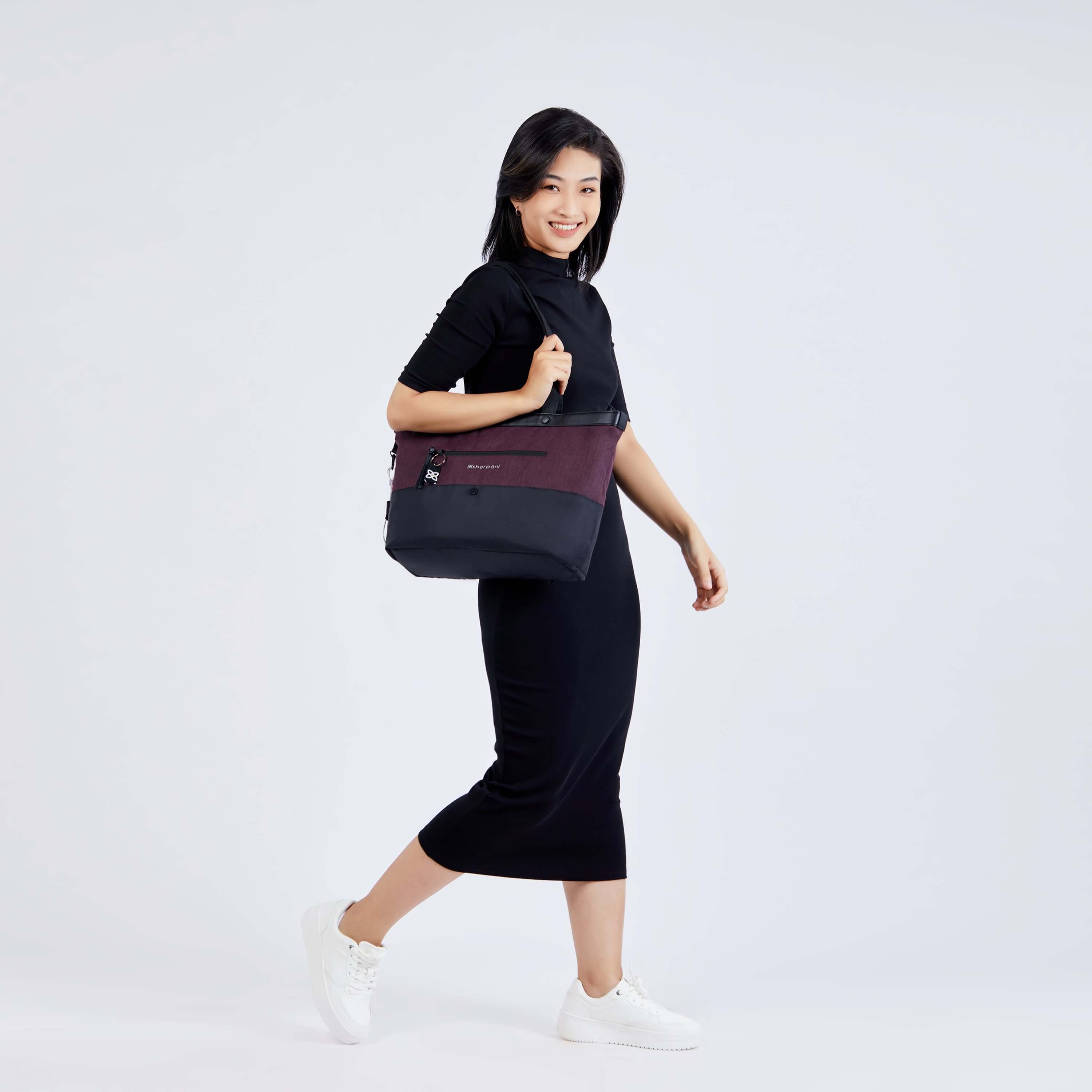 Full body view of a dark hared model facing the side and smiling at the camera. She is wearing a black dress and white shoes. She carries Sherpani&#39;s Anti-Theft tote, the Cali AT in Merlot, on her right shoulder.
