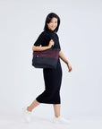 Full body view of a dark hared model facing the side and smiling at the camera. She is wearing a black dress and white shoes. She carries Sherpani's Anti-Theft tote, the Cali AT in Merlot, on her right shoulder.