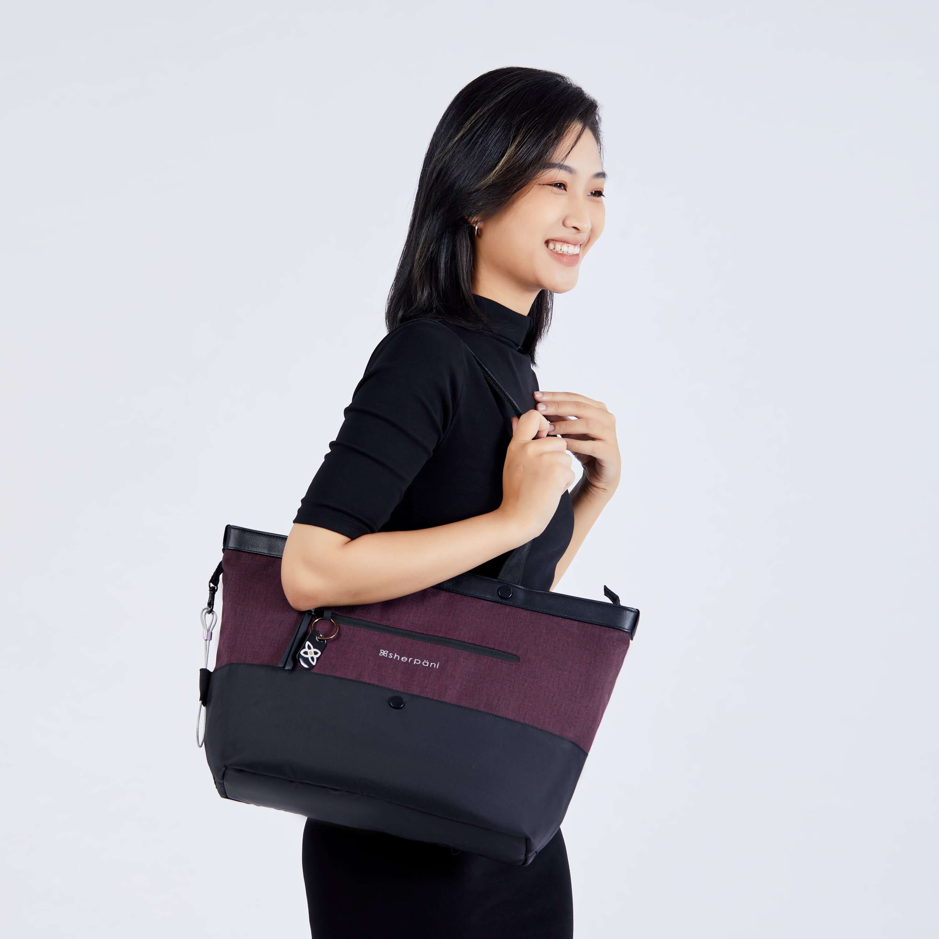 A dark haired model faces the side and smiles. She is wearing a black dress, and is carrying Sherpani's Anti-Theft tote, the Cali AT in Merlot, on her right shoulder. 