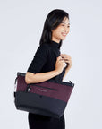 A dark haired model faces the side and smiles. She is wearing a black dress, and is carrying Sherpani's Anti-Theft tote, the Cali AT in Merlot, on her right shoulder.