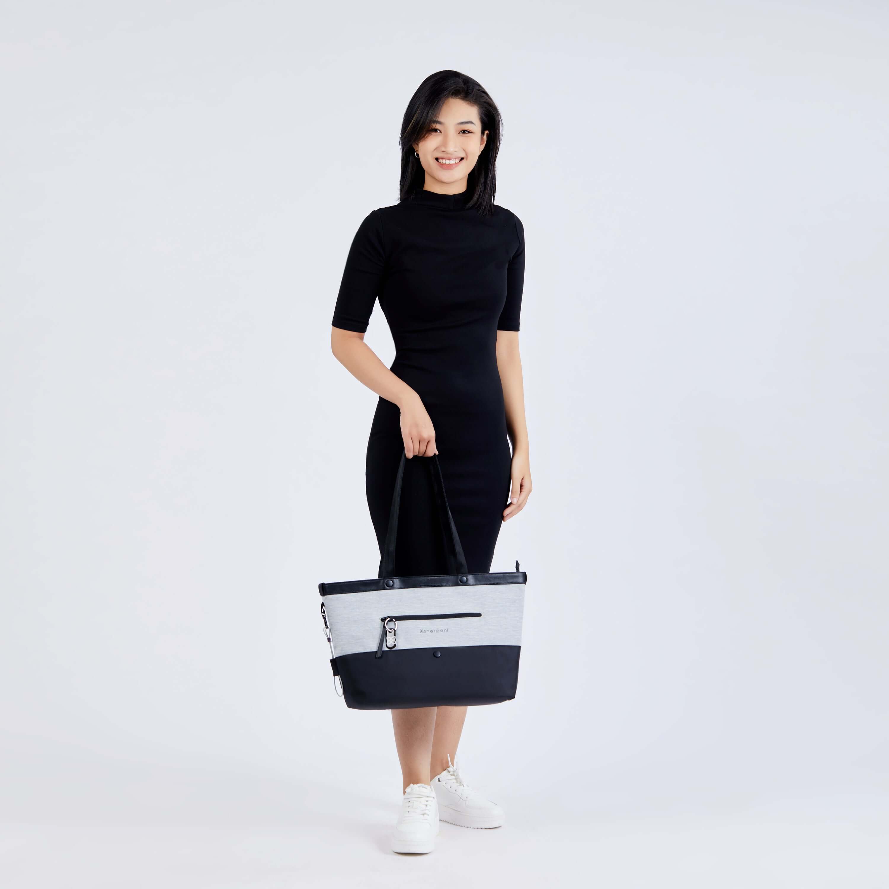 Full body view of a dark haired model facing the camera. She is wearing a black dress and white shoes. She is grabbing the handles of Sherpani's Anti-Theft tote, the Cali AT in Sterling, with her right hand. 