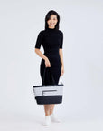 Full body view of a dark haired model facing the camera. She is wearing a black dress and white shoes. She is grabbing the handles of Sherpani's Anti-Theft tote, the Cali AT in Sterling, with her right hand.