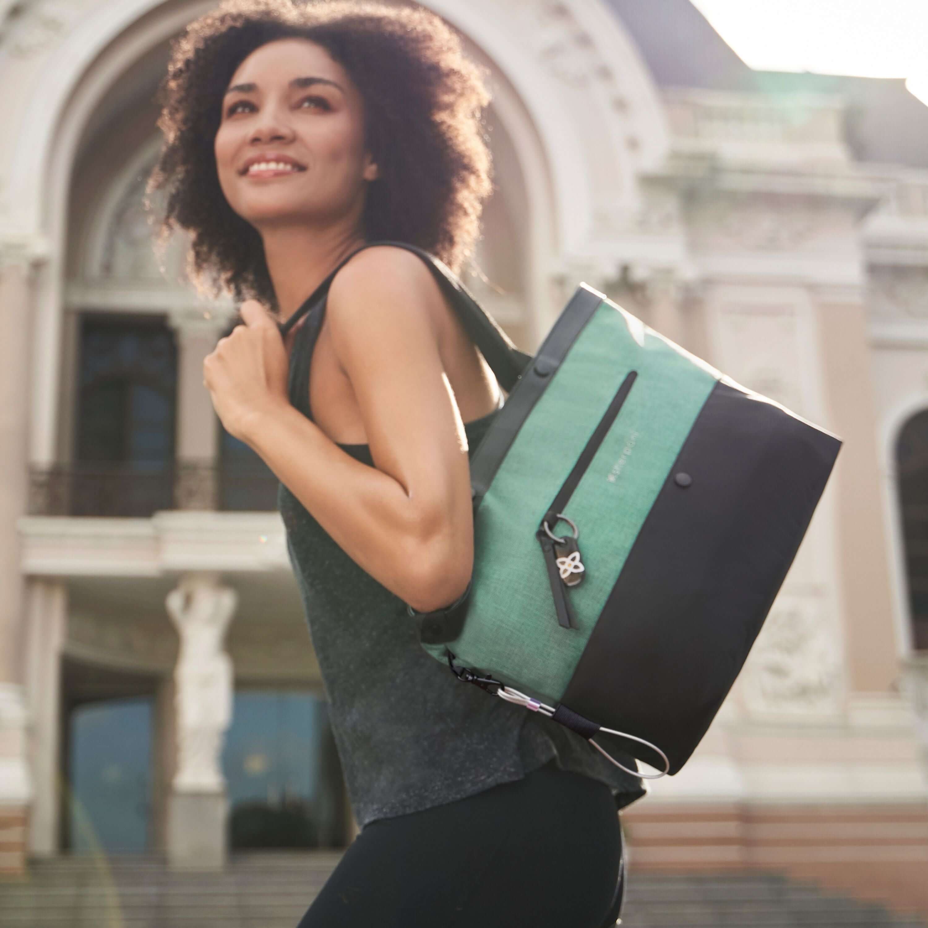 A curly haired model is smiling. She is wearing a gray tank top and black leggings. She is holding Sherpani&#39;s Anti-Theft Tote, the Cali AT in Teal, over her shoulder.