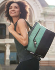 A curly haired model is smiling. She is wearing a gray tank top and black leggings. She is holding Sherpani's Anti-Theft Tote, the Cali AT in Teal, over her shoulder.