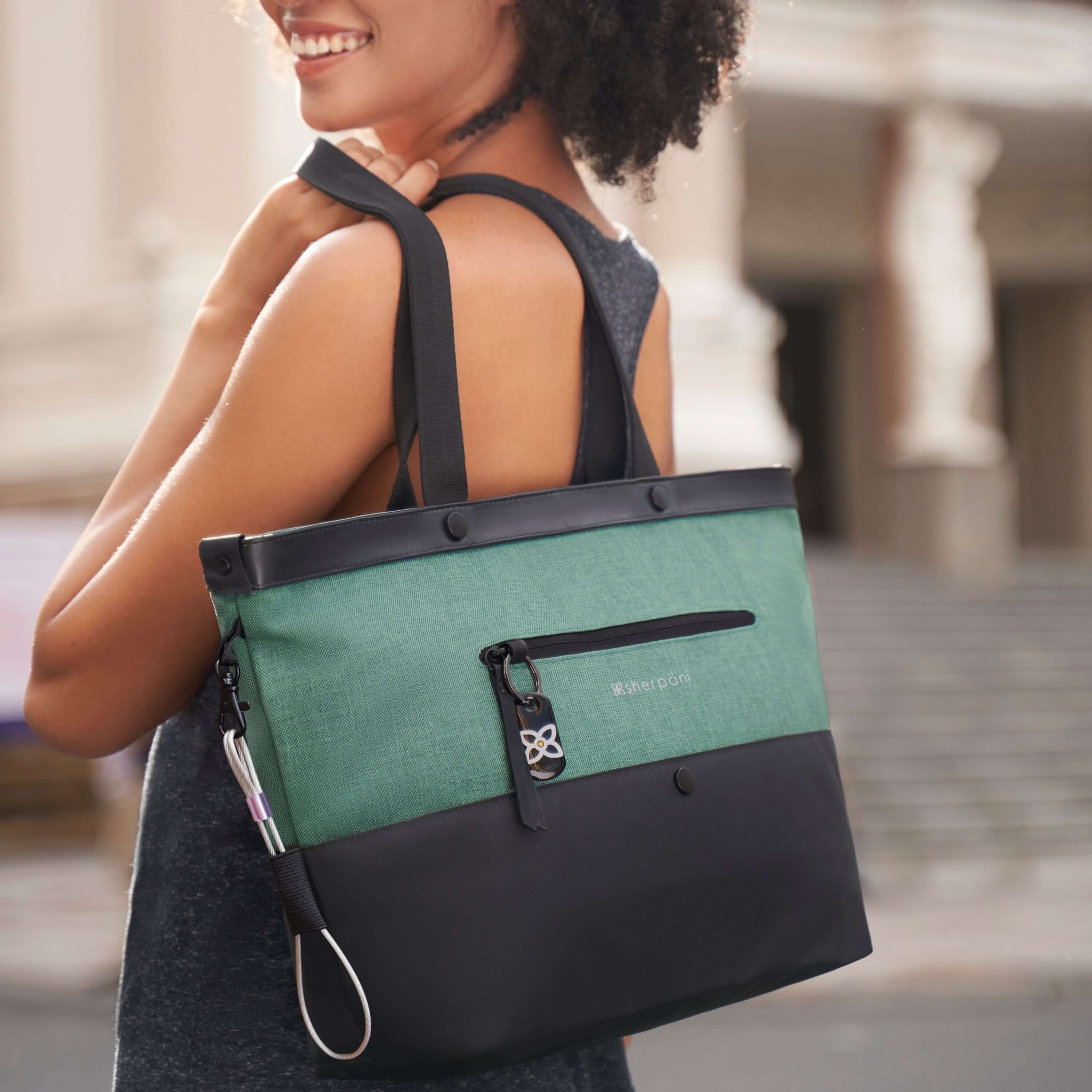 A curly haired model is facing away from the camera, looking over her left shoulder and smiling. She is wearing a gray tank top and black leggings. She is holding Sherpani&#39;s Anti-Theft Tote, the Cali AT in Teal, over her shoulder.