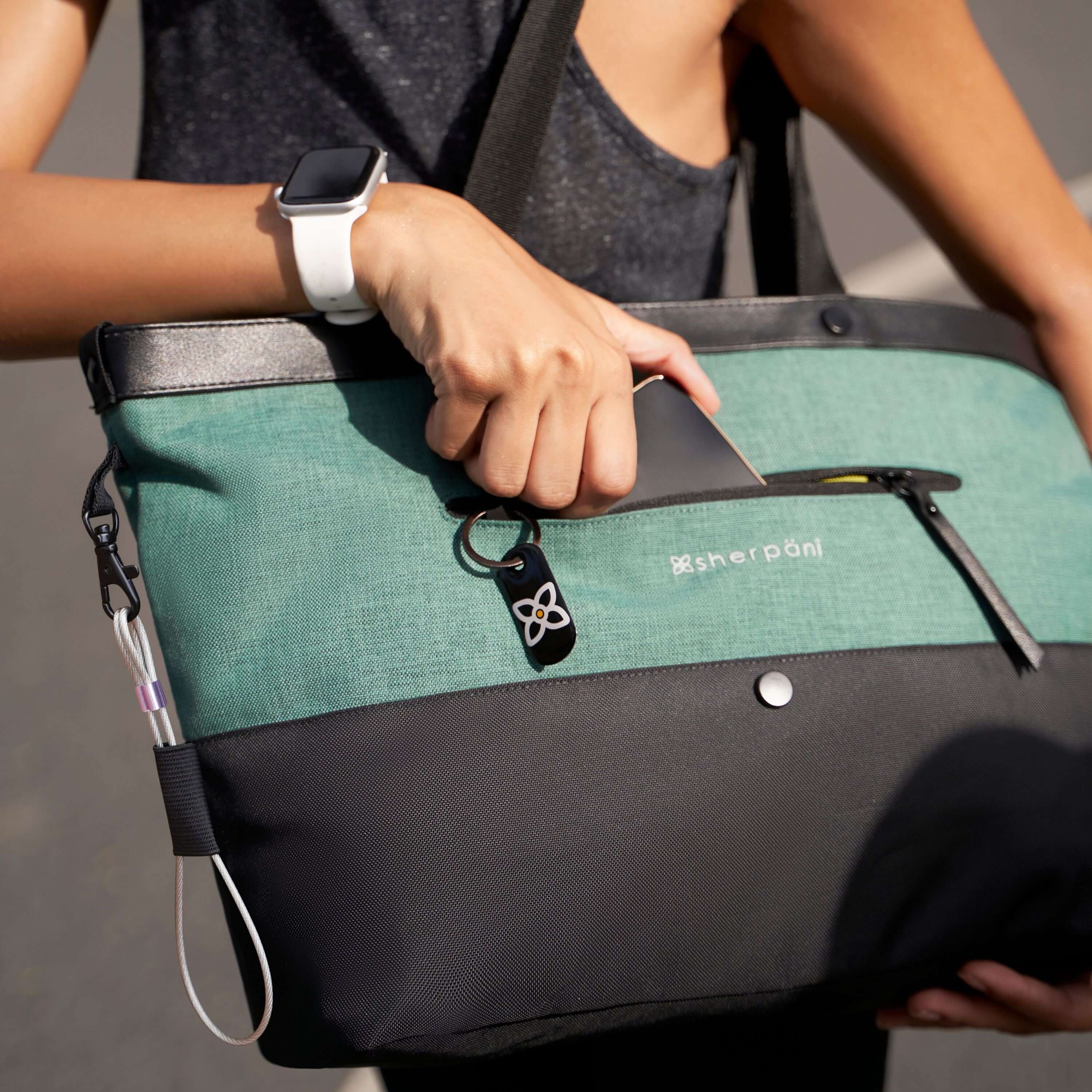 Close up view of a model&#39;s hands putting a phone into the the front pocket of the Sherpani&#39;s Anti-Theft Tote, the Cali AT in Teal.