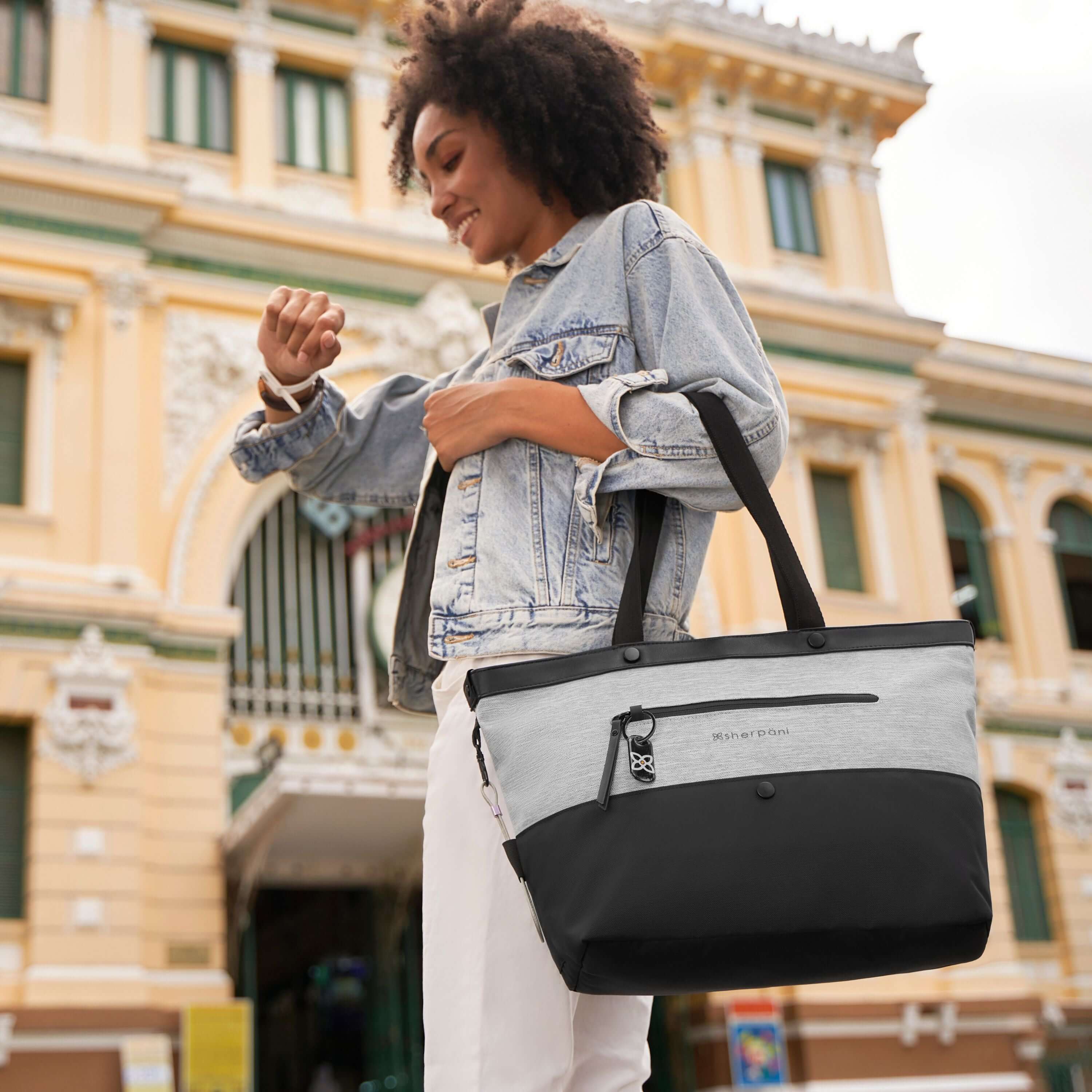 A curly haired model looking at her watch and smiling. She is wearing a jean jacket, white pants, and is carrying Sherpani's Anti-Theft tote the Cali AT in Sterling. 