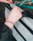 Close up view of a model’s hands demonstrating the zipper lock of the Sherpani's Anti-Theft tote the Cali AT in Sterling. A wire loop lock is hooked around a chair leg. 
