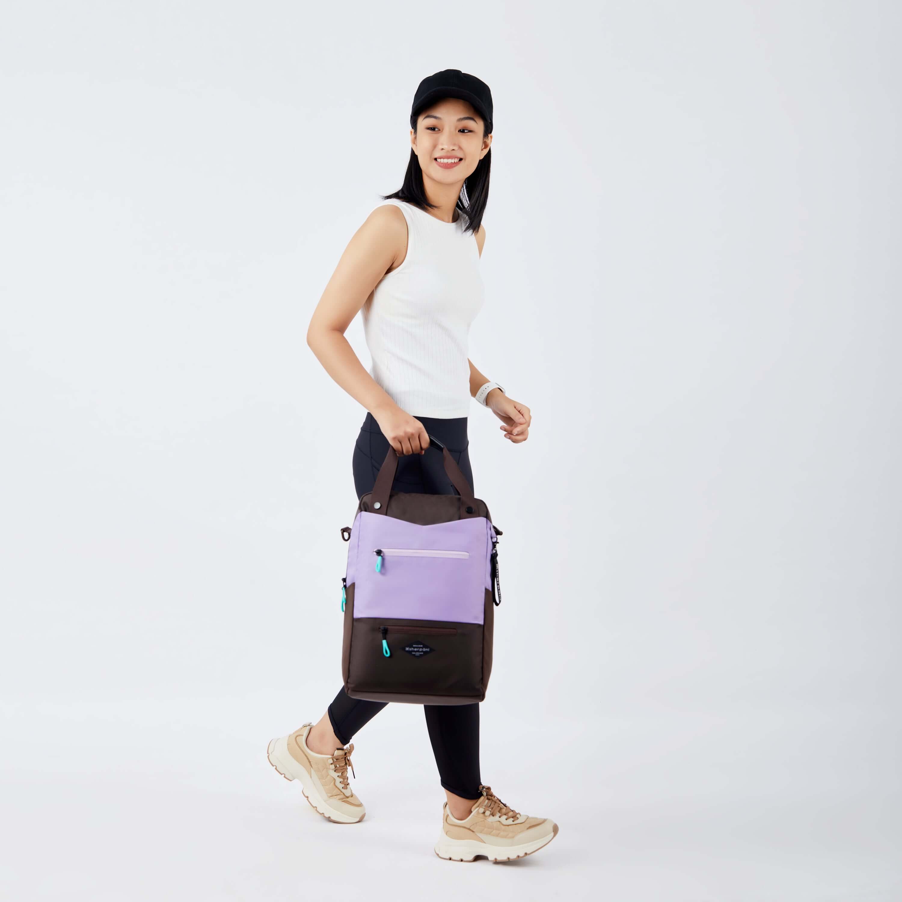 Side view of a dark haired model walking. She is wearing a black hat, white tank top, black leggings and sneakers. She is carrying Sherpani's three-in-one bag, the Camden in Lavender, by the tote handles. 