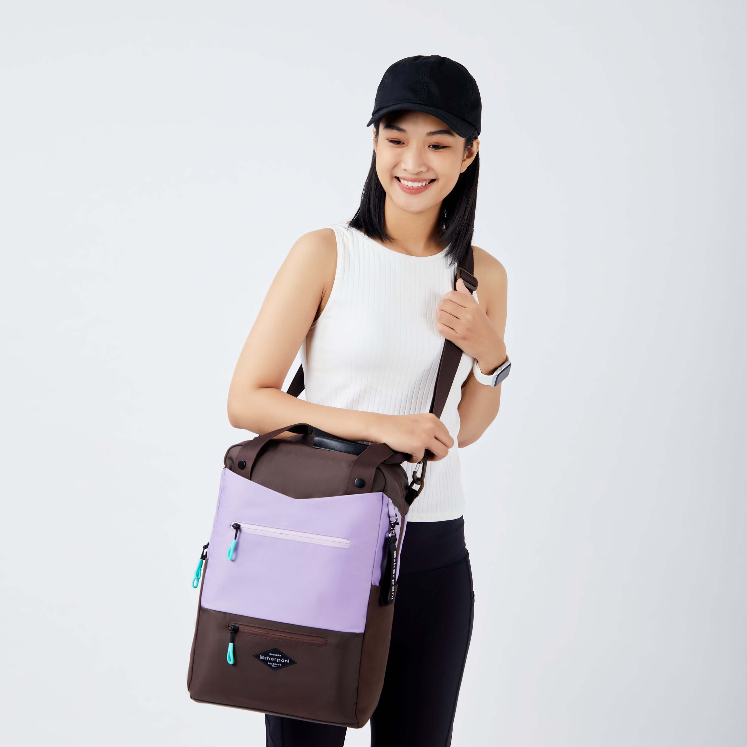 Close up view of a dark haired model facing  the camera and smiling. She is wearing a black hat, white tank top and black leggings. She is carrying Sherpani&#39;s three-in-one bag, the Camden in Lavender, as a crossbody.