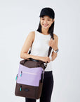 Close up view of a dark haired model facing  the camera and smiling. She is wearing a black hat, white tank top and black leggings. She is carrying Sherpani's three-in-one bag, the Camden in Lavender, as a crossbody.