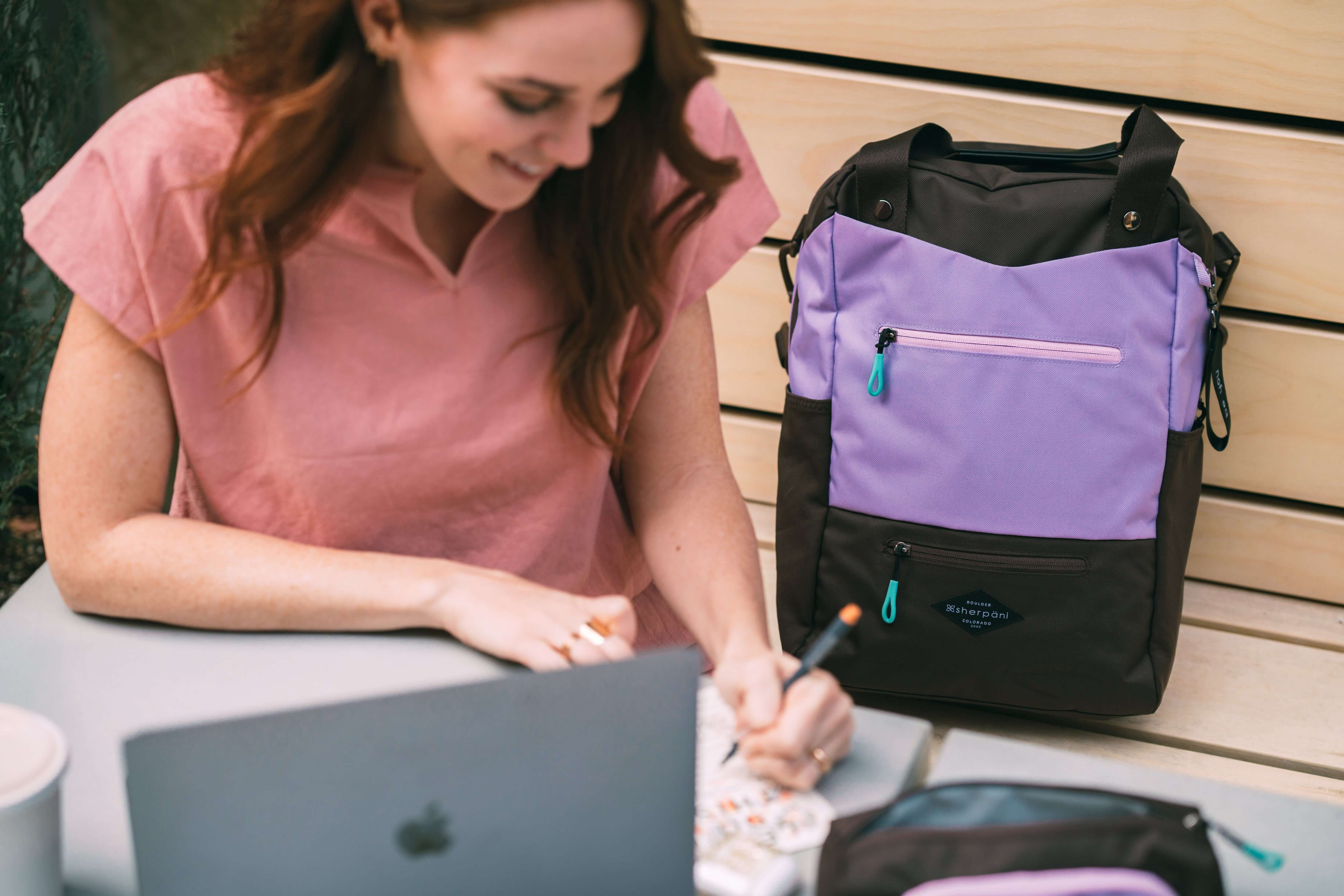 A red haired woman sits at a table outside writing, her laptop and coffee sit in front of her. She is wearing a pink top. Next to her, sits Sherpani&#39;s three-in-one bag, the Camden in Lavender.