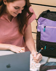 A red haired woman sits at a table outside writing, her laptop and coffee sit in front of her. She is wearing a pink top. Next to her, sits Sherpani's three-in-one bag, the Camden in Lavender.