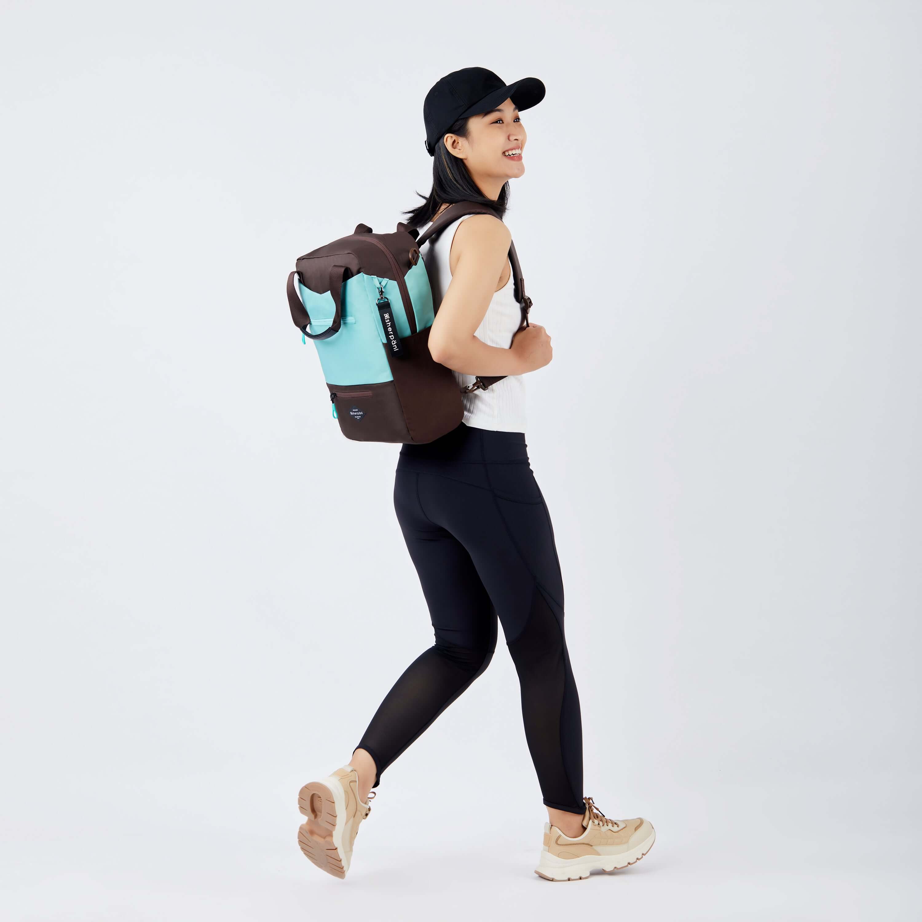 Full body view of a dark haired model walking away from the camera and smiling over her right shoulder. She is wearing a black hat, white tank top, black leggings and sneakers. She is carrying Sherpani&#39;s three-in-one bag, the Camden in Seagreen, as a backpack.