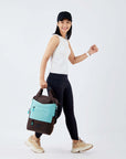 Side view of a dark haired model walking. She is wearing a black hat, white tank top, black leggings and sneakers. She is carrying Sherpani's three-in-one bag, the Camden in Seagreen, by the tote handles.