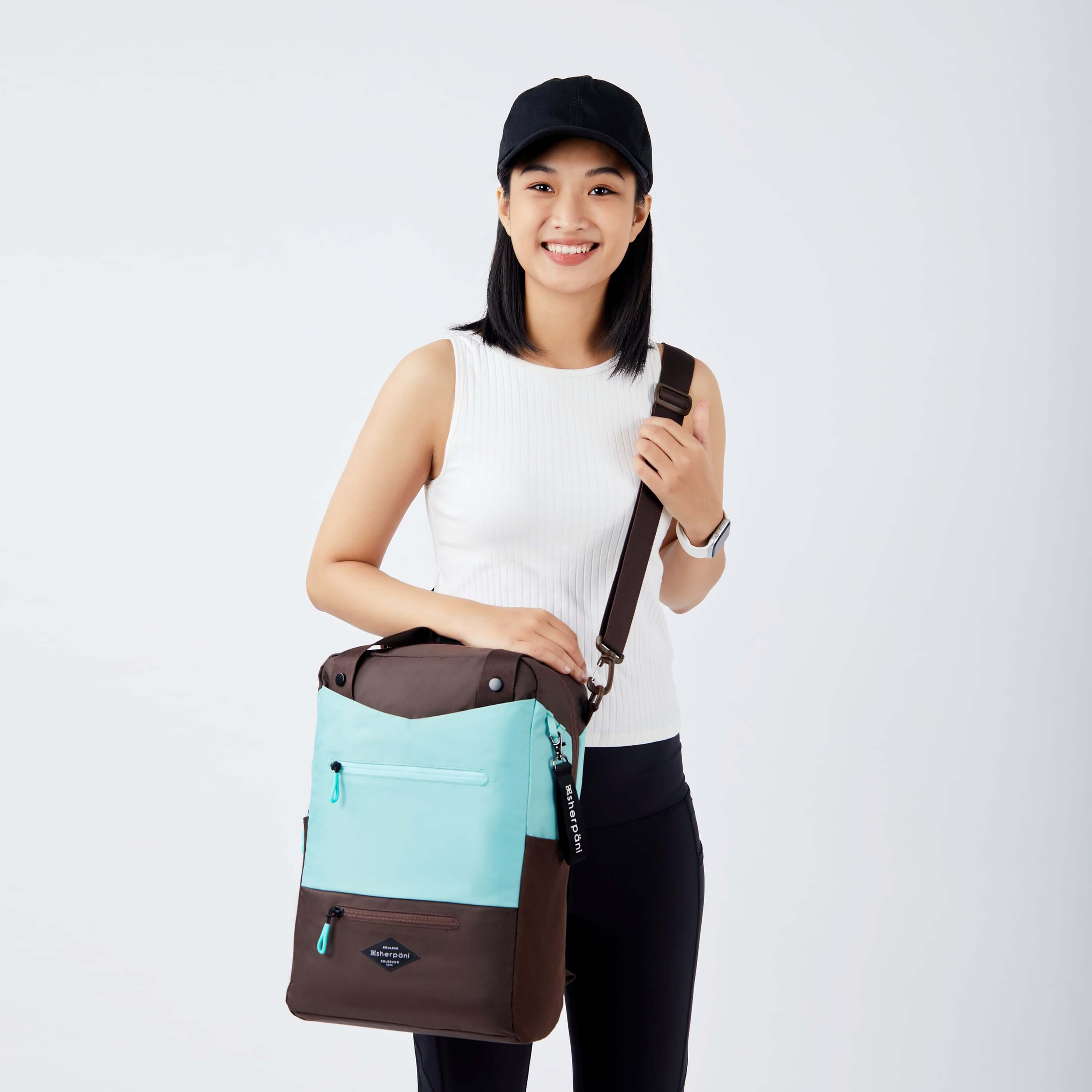 Close up view of a dark haired model smiling at the camera. She is wearing a black hat, white tank top and black leggings. She is carrying Sherpani's three-in-one bag, the Camden in Seagreen, as a crossbody. 