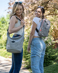Two smiling women stand side-by-side on an outdoor path in front of a blossoming tree. The woman on the left has blonde hair, she is wearing a white tank top and jeans. She carries Sherpani mesh crossbody, the Payton. The woman on the right has brown hair, she is wearing a white tee shirt and jeans. She carries Sherpani mesh backpack, the Adaline.