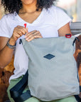 A curly haired woman sits on a bench outdoors. She is wearing a white tee shirt and green pants. She is unzipping the main compartment of Sherpani mesh convertible bag, the Delanie.