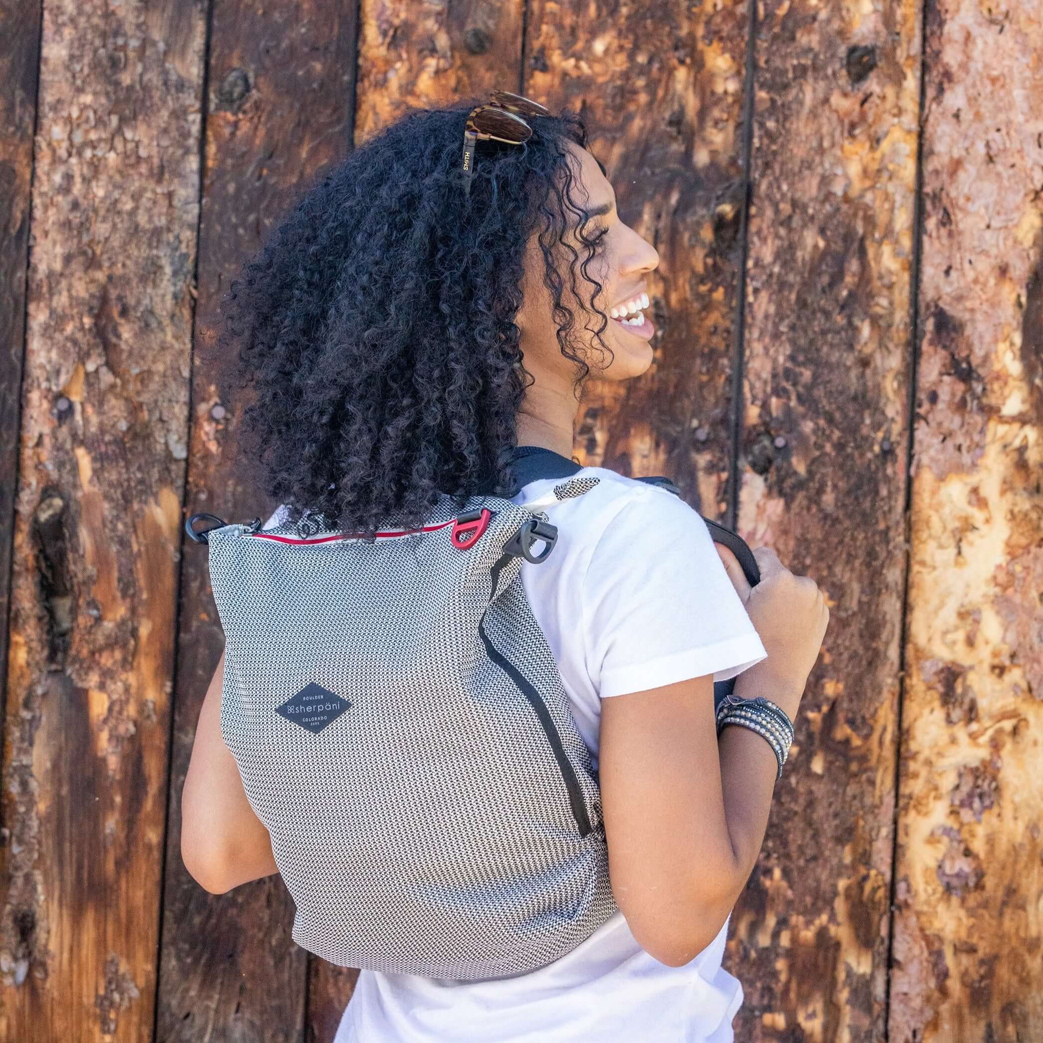 A curly haired woman stands outside in front of a wooden wall. She is facing away from the camera and smiling over her right shoulder. She is wearing a white tee shirt. She carries Sherpani mesh convertible bag, the Delanie, as a backpack.