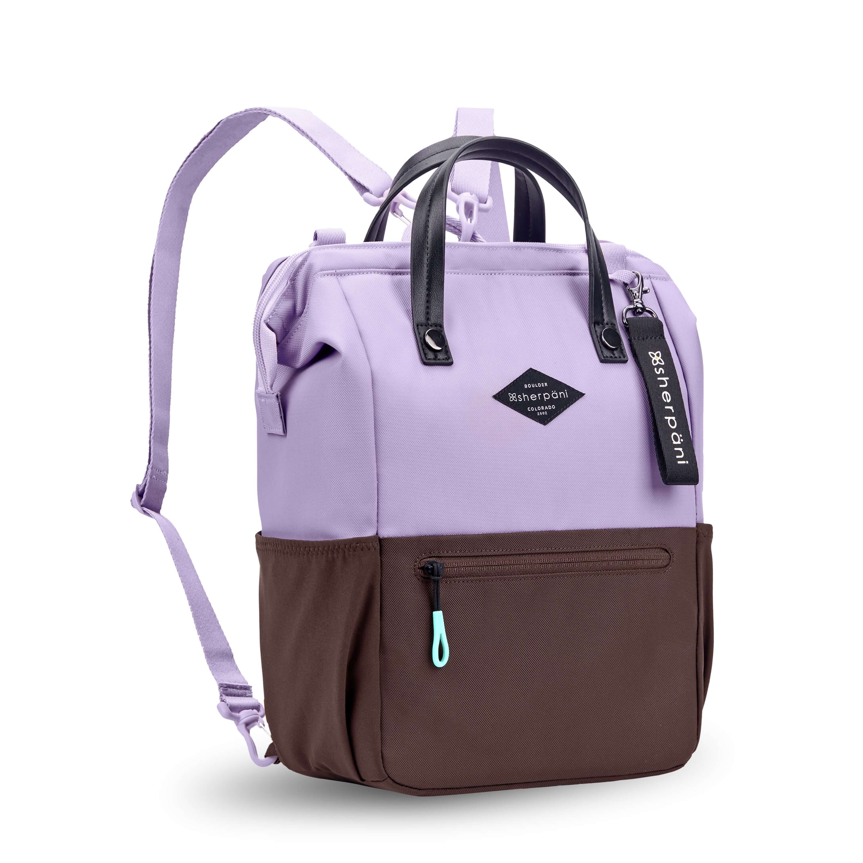 Angled front view of Sherpani three-in-one bag, the Dispatch in Lavender. The bag is two-toned: the top is lavender and the bottom is brown. There is an external zipper pocket on the front panel. Easy-pull zippers are accented in aqua. A branded Sherpani keychain is clipped to the upper right corner. Elastic water bottle holders sit on either side of the bag. It has short tote handles and adjustable/detachable straps that can function for a backpack or crossbody.