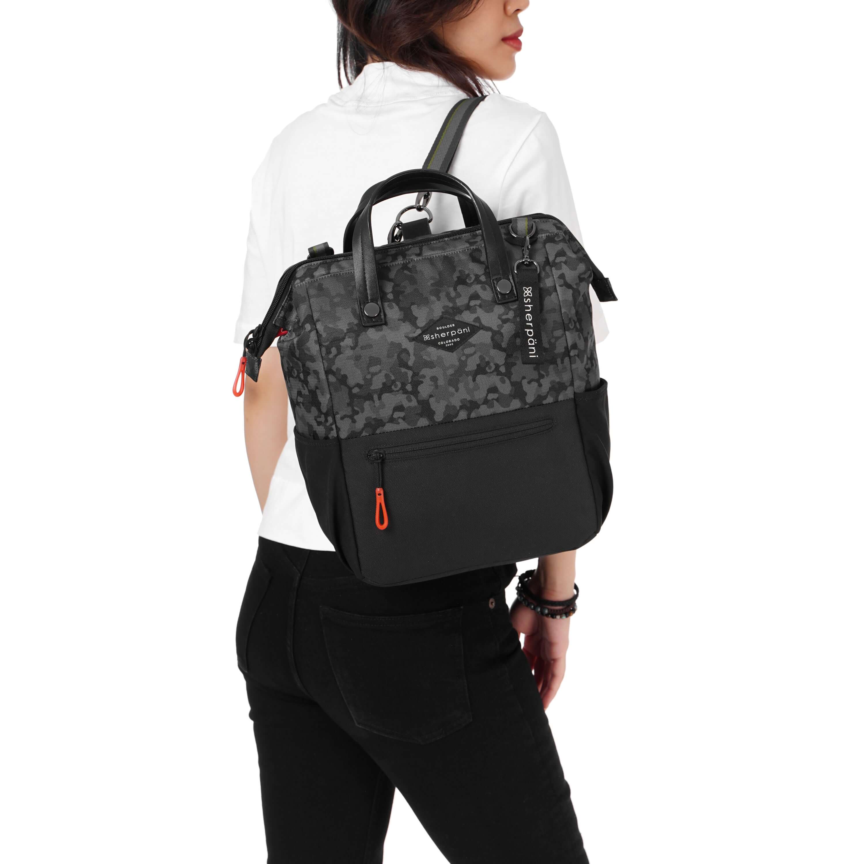 Close up view of a dark haired model facing away from the camera and looking over her right shoulder. She is wearing a white tee shirt and black pants. She carries Sherpani three-in-one bag, the Dispatch in Dream Camo, as a backpack.