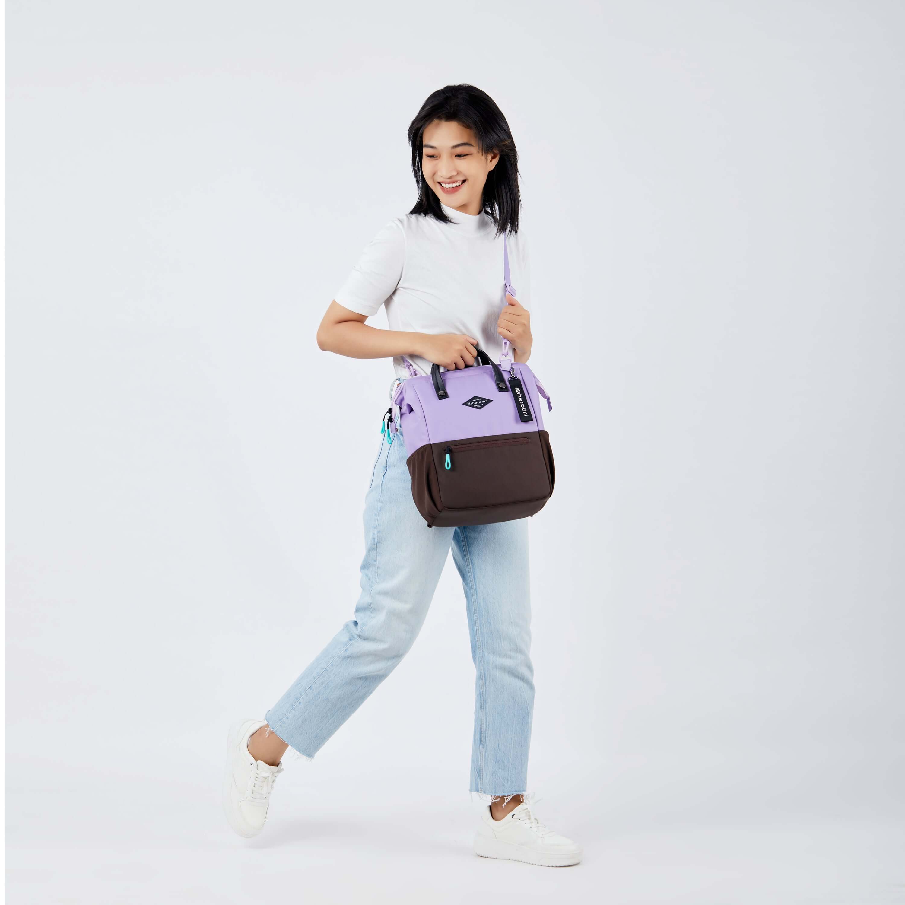 Full body view of a dark haired model walking to the side. She is wearing a white tee shirt, jeans and white sneakers. She carries Sherpani three-in-one bag, the Dispatch in Lavender, as a crossbody.