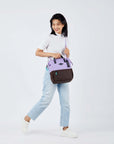 Full body view of a dark haired model walking to the side. She is wearing a white tee shirt, jeans and white sneakers. She carries Sherpani three-in-one bag, the Dispatch in Lavender, as a crossbody.