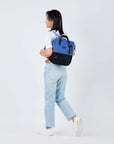 Full body view of a dark haired model facing away from the camera and walking. She is wearing a white tee shirt, jeans and white sneakers. She carries Sherpani three-in-one bag, the Dispatch in Pacific Blue, as a backpack.