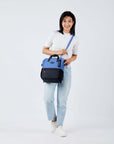 Full body view of a dark haired model facing the camera and smiling. She is wearing a white tee shirt, jeans and white sneakers. She carries Sherpani three-in-one bag, the Dispatch in Pacific Blue, as a crossbody.