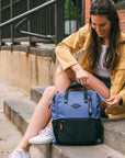 A brown haired woman sits on a cement staircase outside. She is wearing a yellow jacket, shorts and white sneakers. She is unzipping the main compartment of Sherpani three-in-one bag, the Dispatch in Pacific Blue.