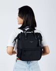 Close up view of a dark haired model facing away from the camera and looking over her left shoulder. She is wearing a white tee shirt and jeans. She carries Sherpani three-in-one bag, the Dispatch in Raven, as a backpack.