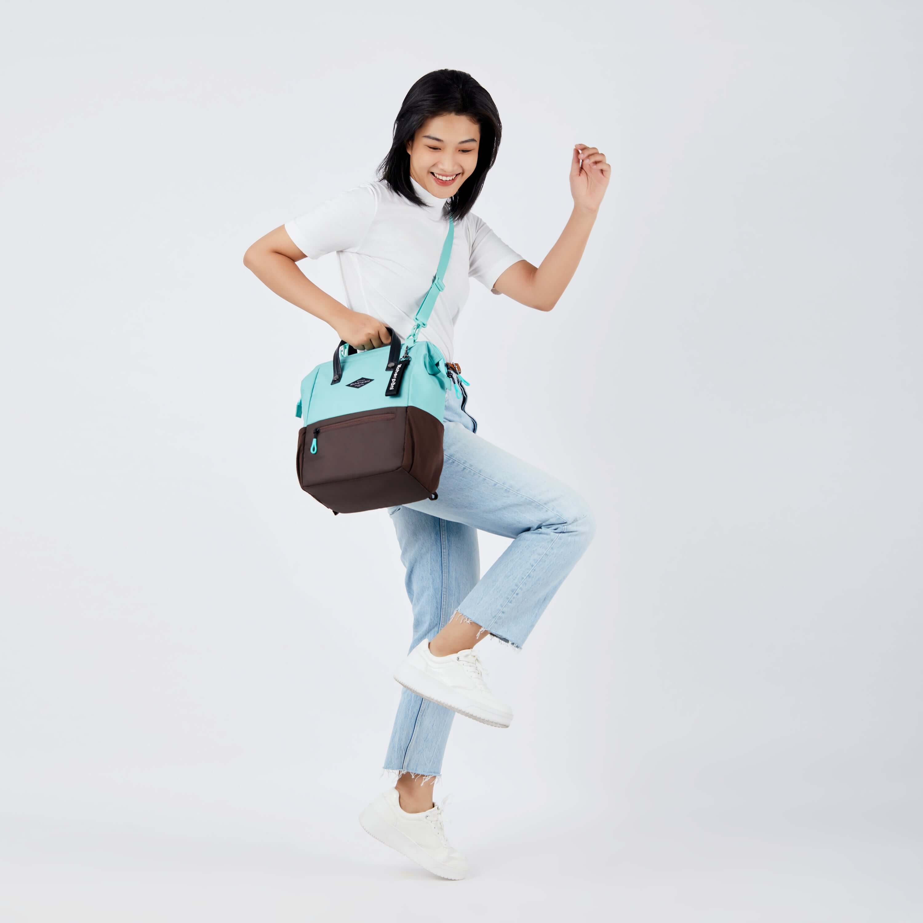 Full body view of a dark haired model skipping to the side and smiling. She is wearing a white tee shirt, jeans and white sneakers. She carries Sherpani three-in-one bag, the Dispatch in Seagreen, as a crossbody. 
