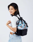Close up view of a dark haired model facing away from the camera and smiling over her left shoulder. She is wearing a white tee shirt and jeans. She carries Sherpani three-in-one bag, the Dispatch in Summer Camo, as a backpack.