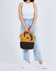 Full body view of a dark haired model facing the camera. She is wearing a white tank top, jeans and white sneakers. She carries Sherpani three-in-one bag, the Dispatch in Sundial, by its tote handles.