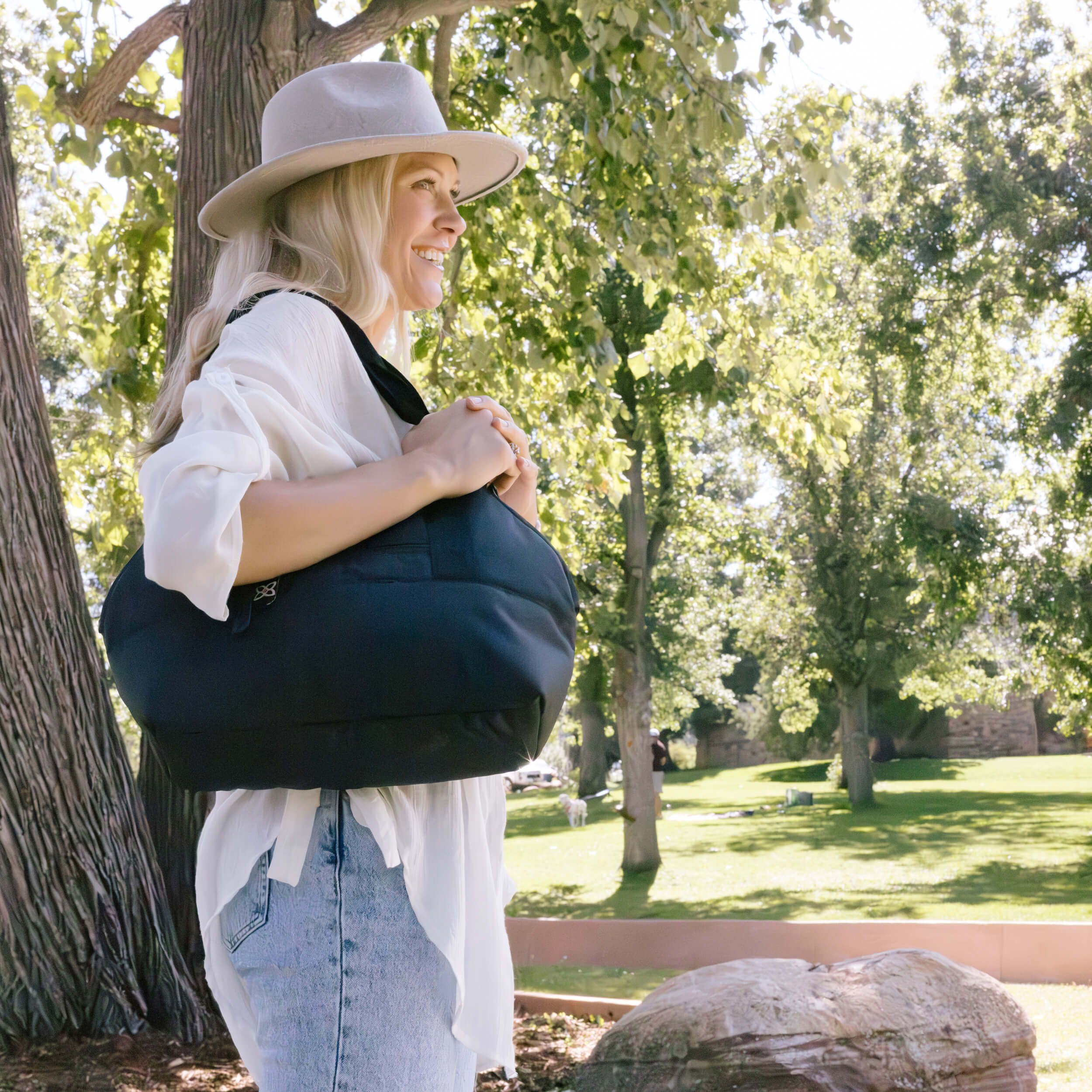A woman standing outdoors in a park wearing the Eclipse in Carbon as a shoulder bag. 