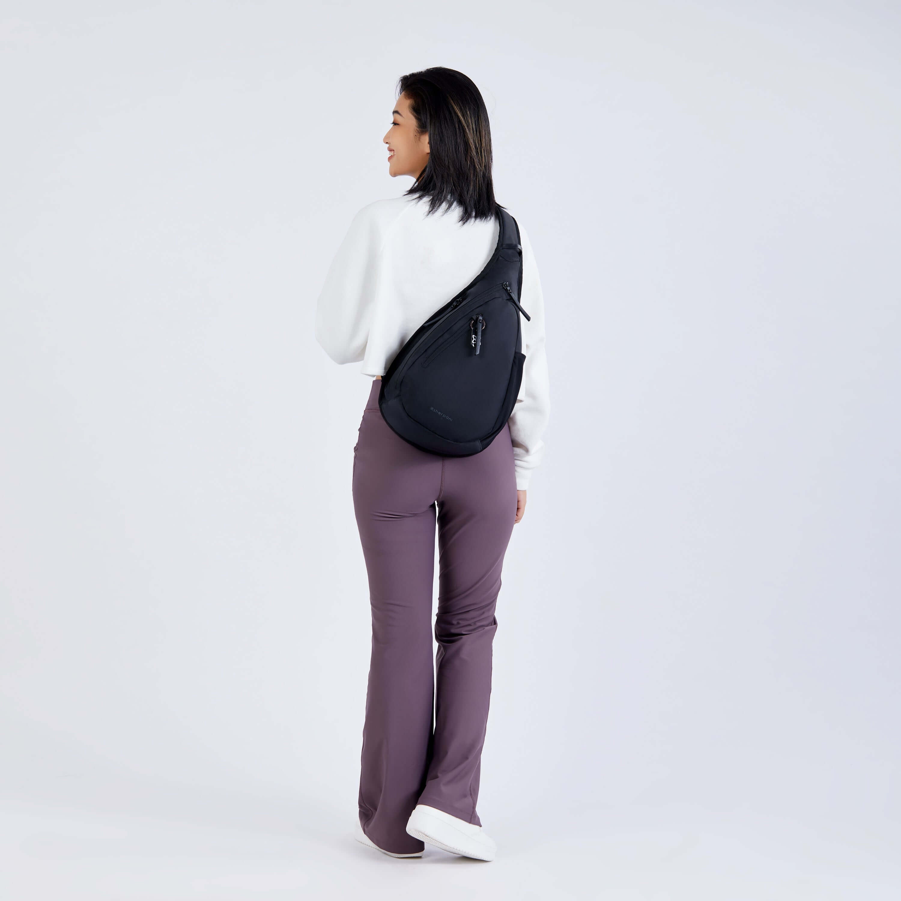 Full body view of a dark haired model facing away from the camera and smiling over her left shoulder. She is wearing a white sweatshirt, purple leggings and Sherpani’s Anti-Theft bag, the Esprit AT in Carbon, across her back.