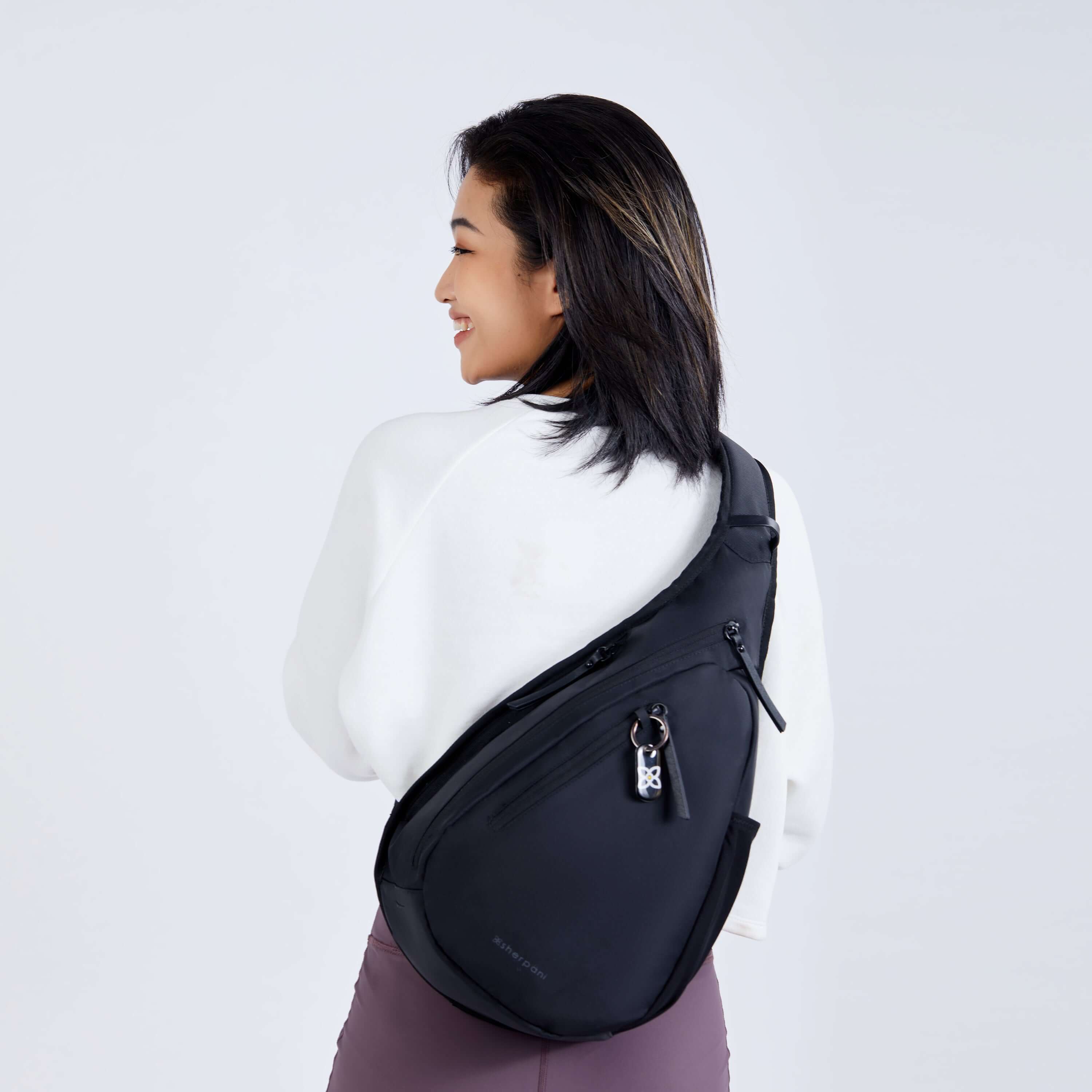 Close up view of a dark haired model facing away from the camera and smiling over her left shoulder. She is wearing a white sweatshirt, purple leggings and Sherpani’s Anti-Theft bag, the Esprit AT in Carbon, across her back. 