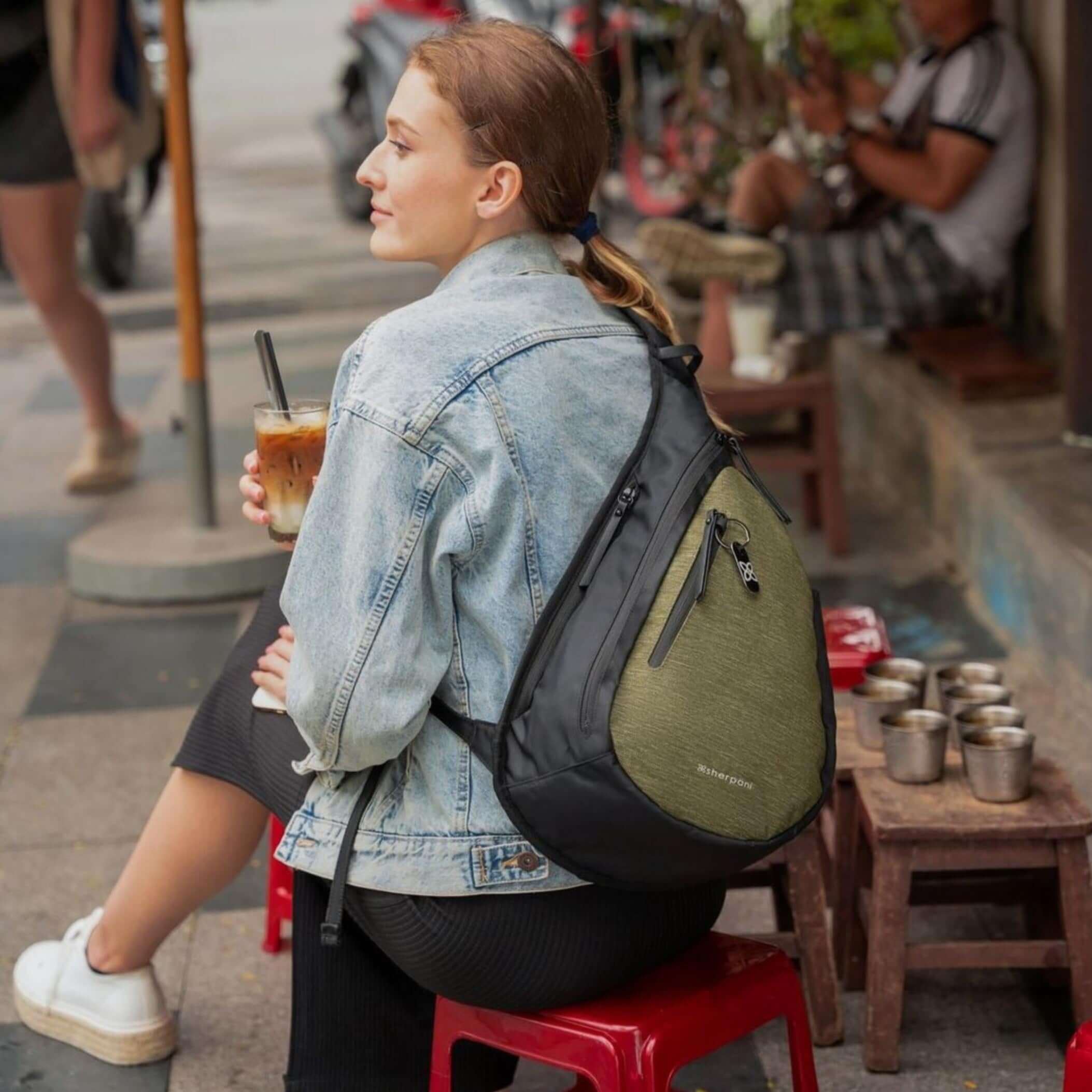 Full body view of a brown haired model sitting at a cafe table outside with an iced coffee. She is facing away from the camera and looking over her left shoulder. She wears a jean jacket, a black skirt, and Sherpani’s Anti-Theft bag, the Esprit AT in Loden, across her back.