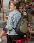 Full body view of a brown haired model sitting at a cafe table outside with an iced coffee. She is facing away from the camera and looking over her left shoulder. She wears a jean jacket, a black skirt, and Sherpani’s Anti-Theft bag, the Esprit AT in Loden, across her back.