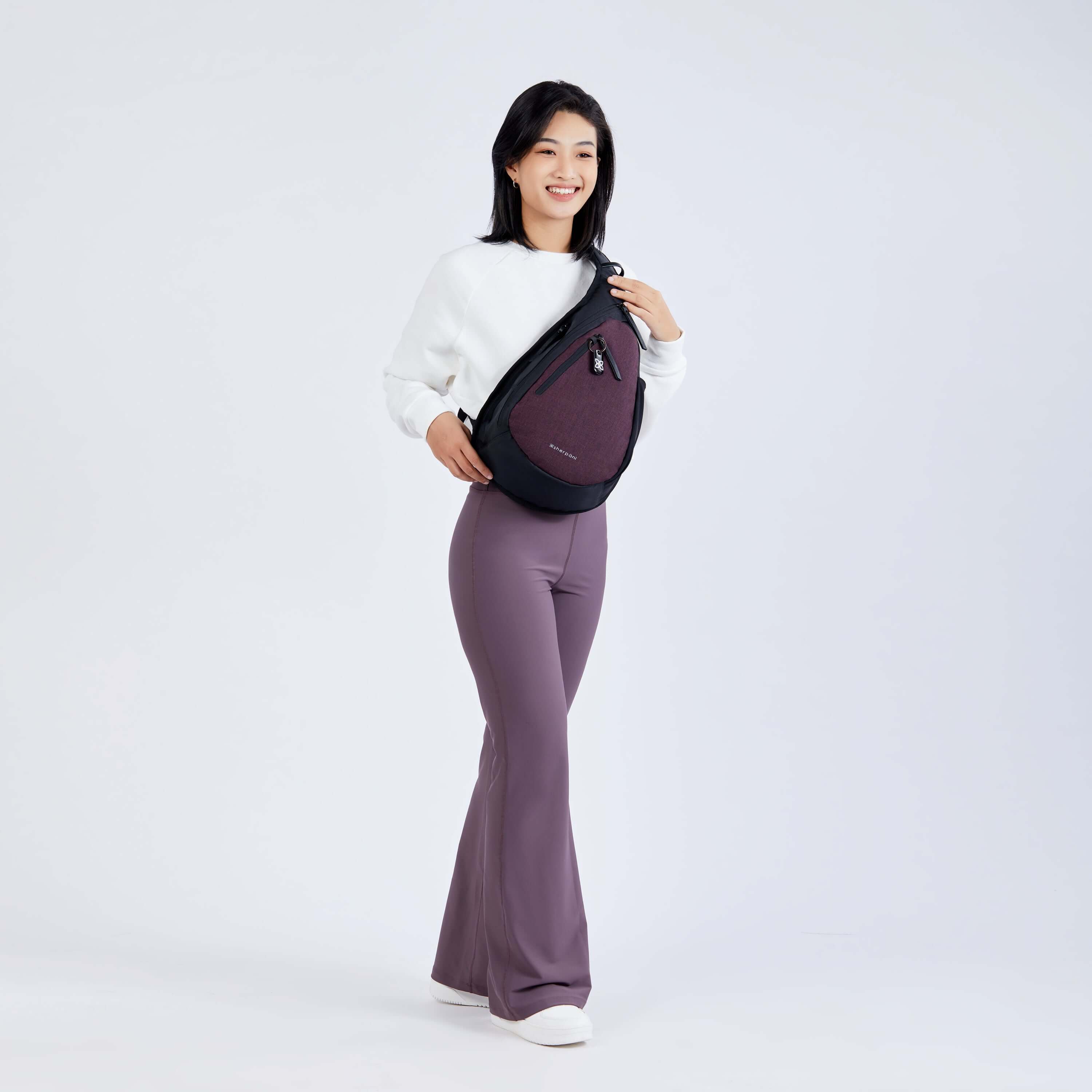 Full body view of a dark haired model facing the camera and smiling. She is wearing a white sweatshirt, purple leggings and Sherpani’s Anti-Theft bag, the Esprit AT in Merlot, as a crossbody. 