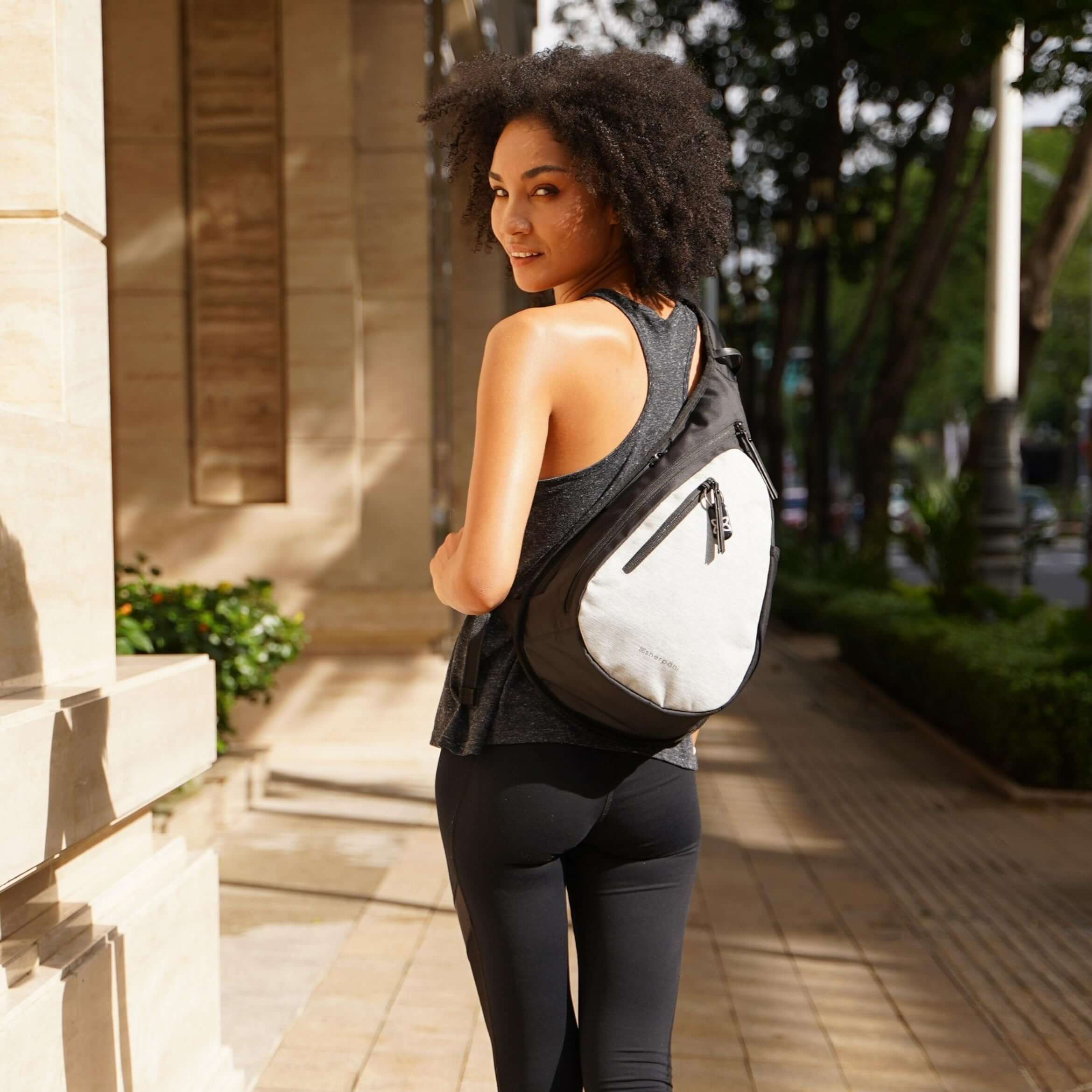 Close up view of a curly haired model facing away from the camera and smiling over her left shoulder. She is wearing a gray tank top, black leggings and Sherpani’s Anti-Theft bag, the Esprit AT in Sterling, across her back.