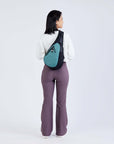 Full body view of a dark haired model facing away from the camera and smiling over her right shoulder. She is wearing a white sweatshirt, purple leggings and Sherpani’s Anti-Theft bag, the Esprit AT in Teal, across her back.