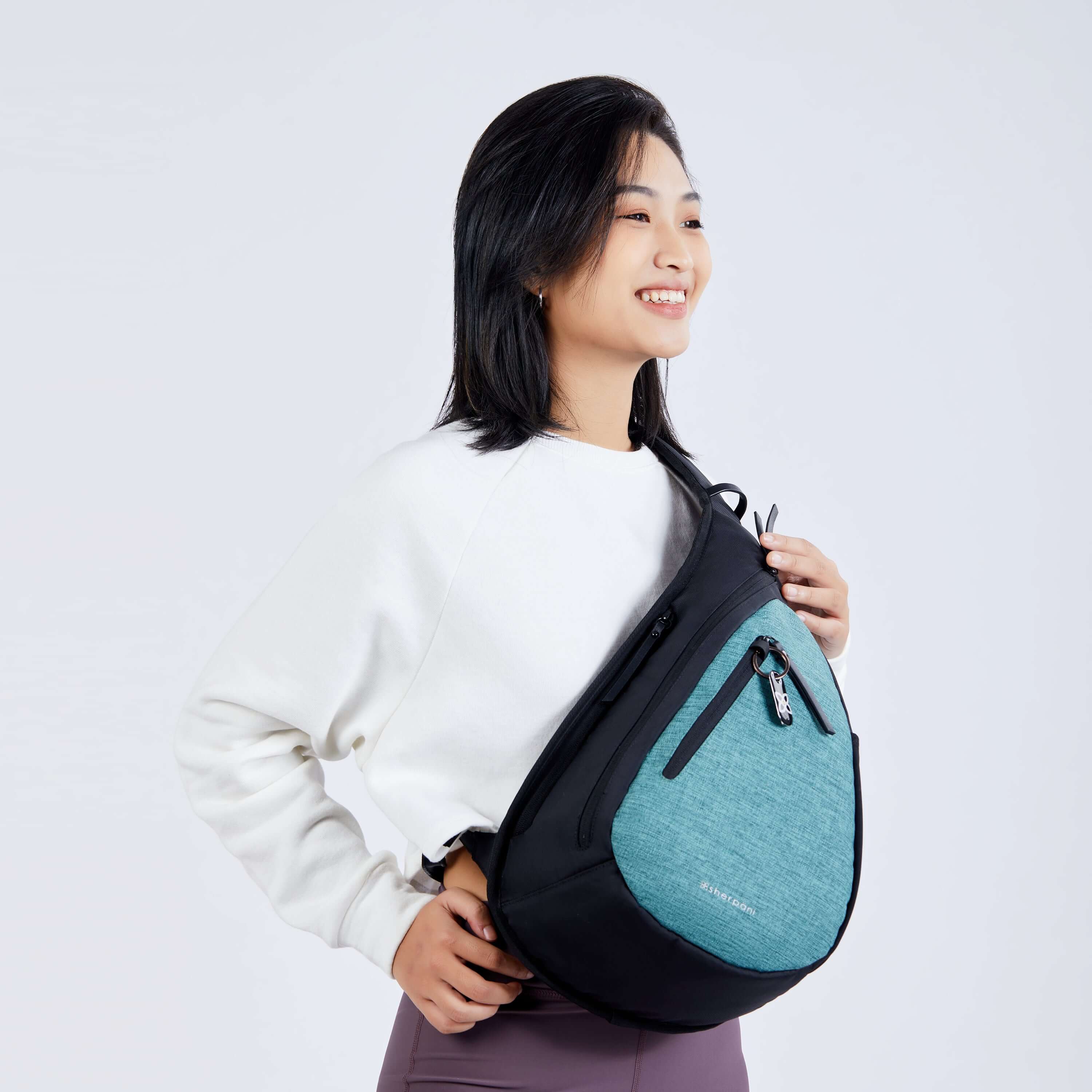 Close up view of a dark haired model facing the camera and smiling. She is wearing a white sweatshirt, purple leggings and Sherpani’s Anti-Theft bag, the Esprit AT in Teal, as a crossbody. 