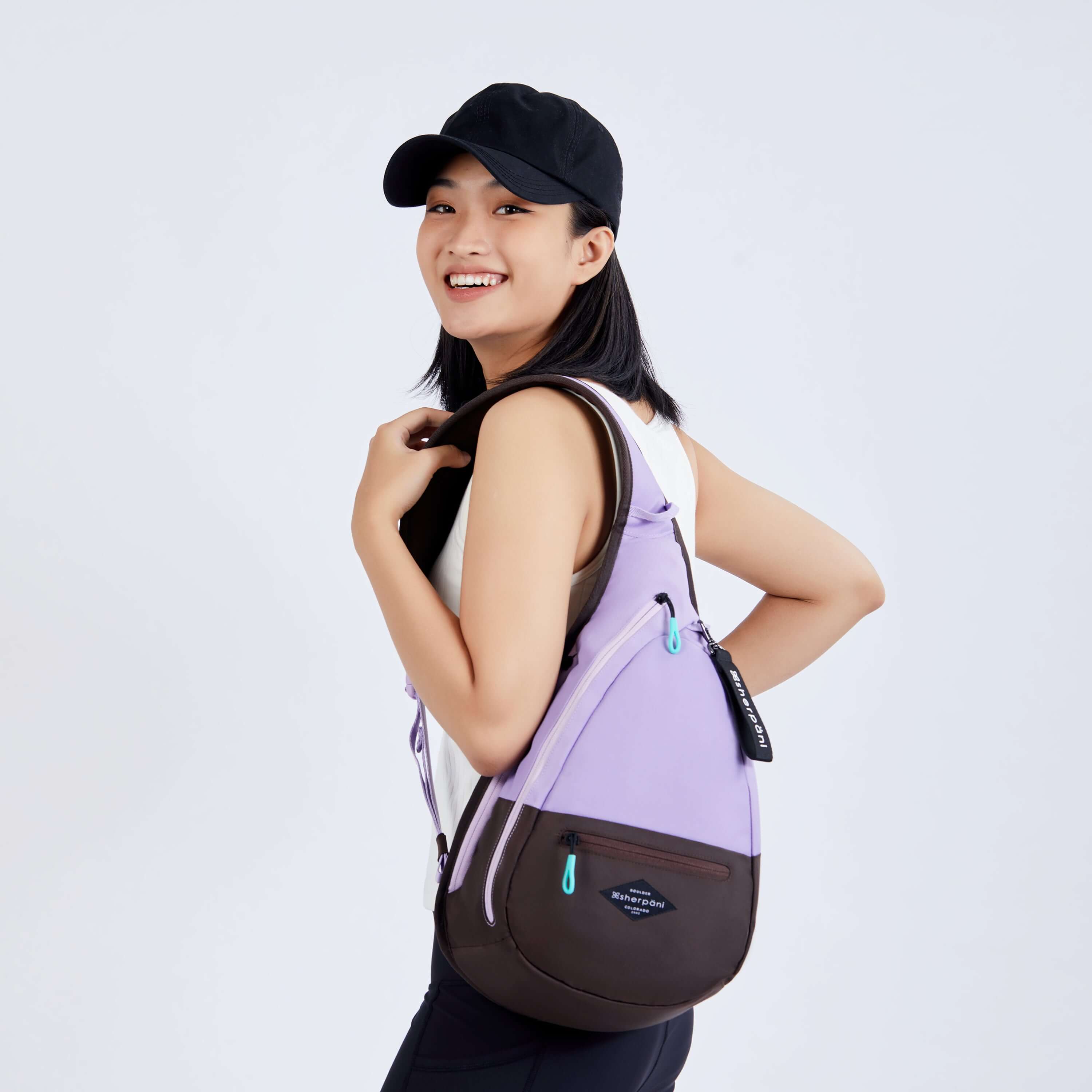 Close up view of a dark haired model smiling over her left shoulder at the camera. She is wearing a black ball cap, white tank top and black leggings. She carries Sherpani bag, the Esprit in Lavender, over one shoulder. 