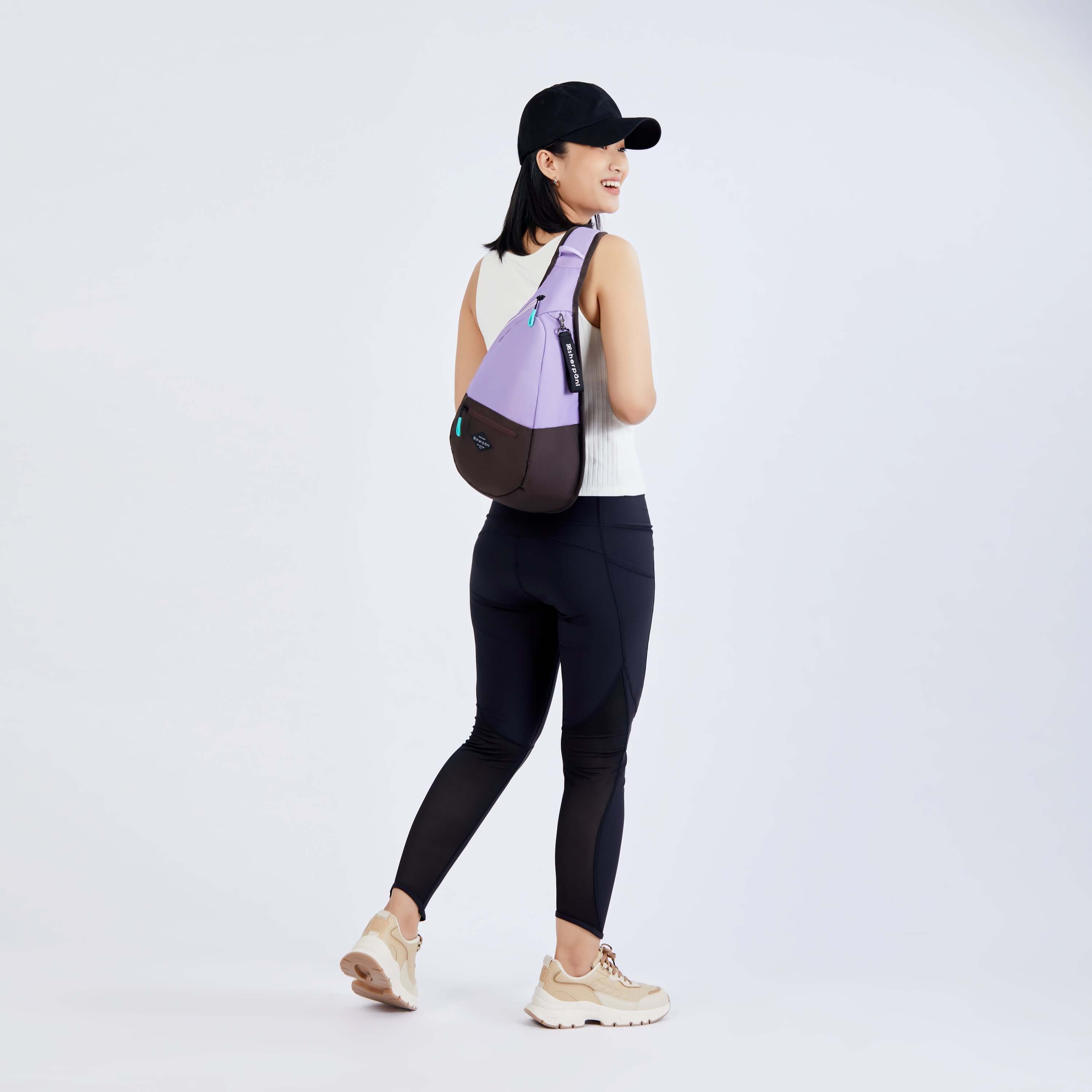 Full body view of a dark haired model facing away from the camera and smiling over her right shoulder. She is wearing a black ball cap, white tank top, black leggings and sneakers. She carries Sherpani bag, the Esprit in Lavender, as a crossbody. 