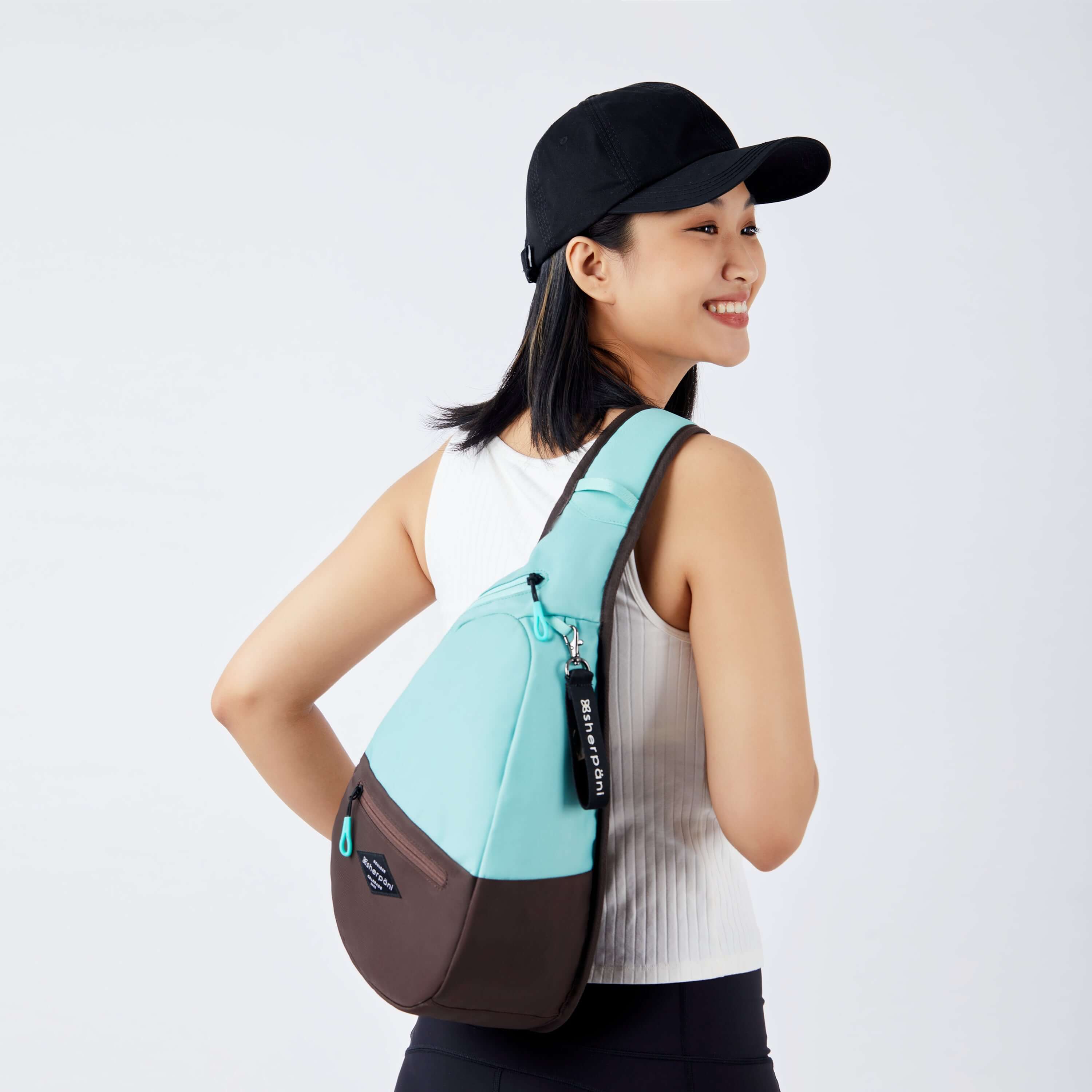 Close up view of a dark haired model facing away from the camera and smiling over her right shoulder. She is wearing a black ball cap, white tank top and black leggings. She carries Sherpani bag, the Esprit in Seagreen, as a crossbody. 