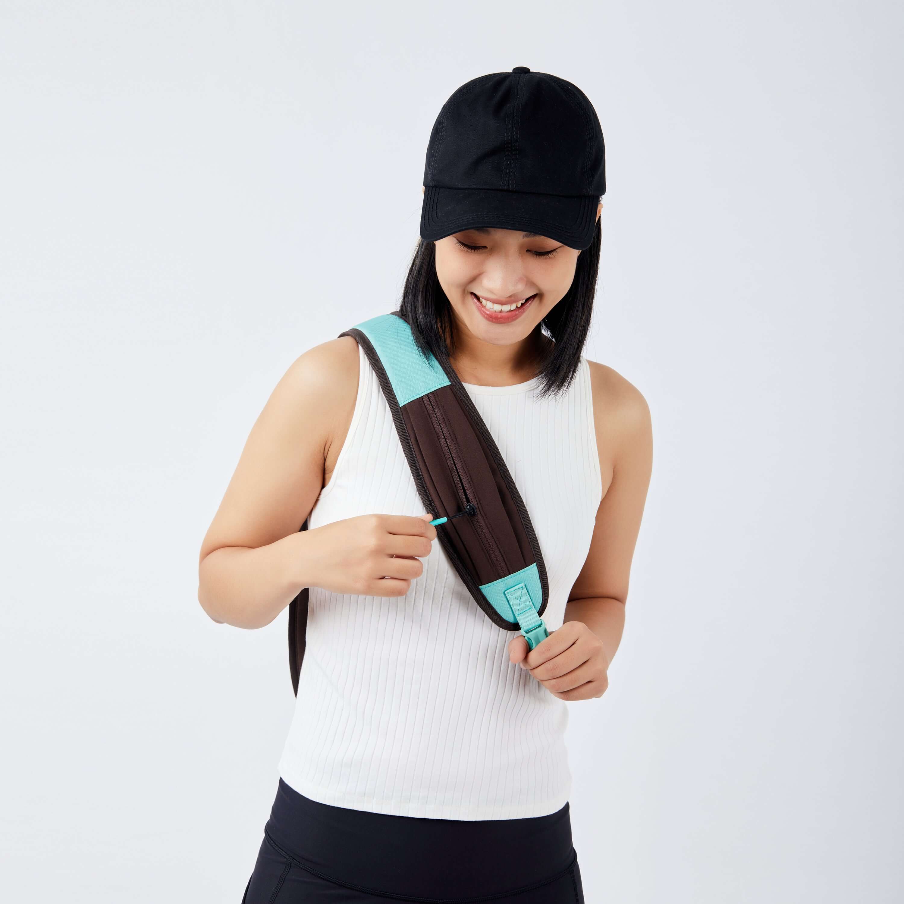 Close up view of a dark haired model facing the camera and smiling downward. She is wearing a black ball cap, white tank top and black leggings. She is opening the small zipper compartment on the crossbody strap of Sherpani bag, the Esprit in Seagreen. 