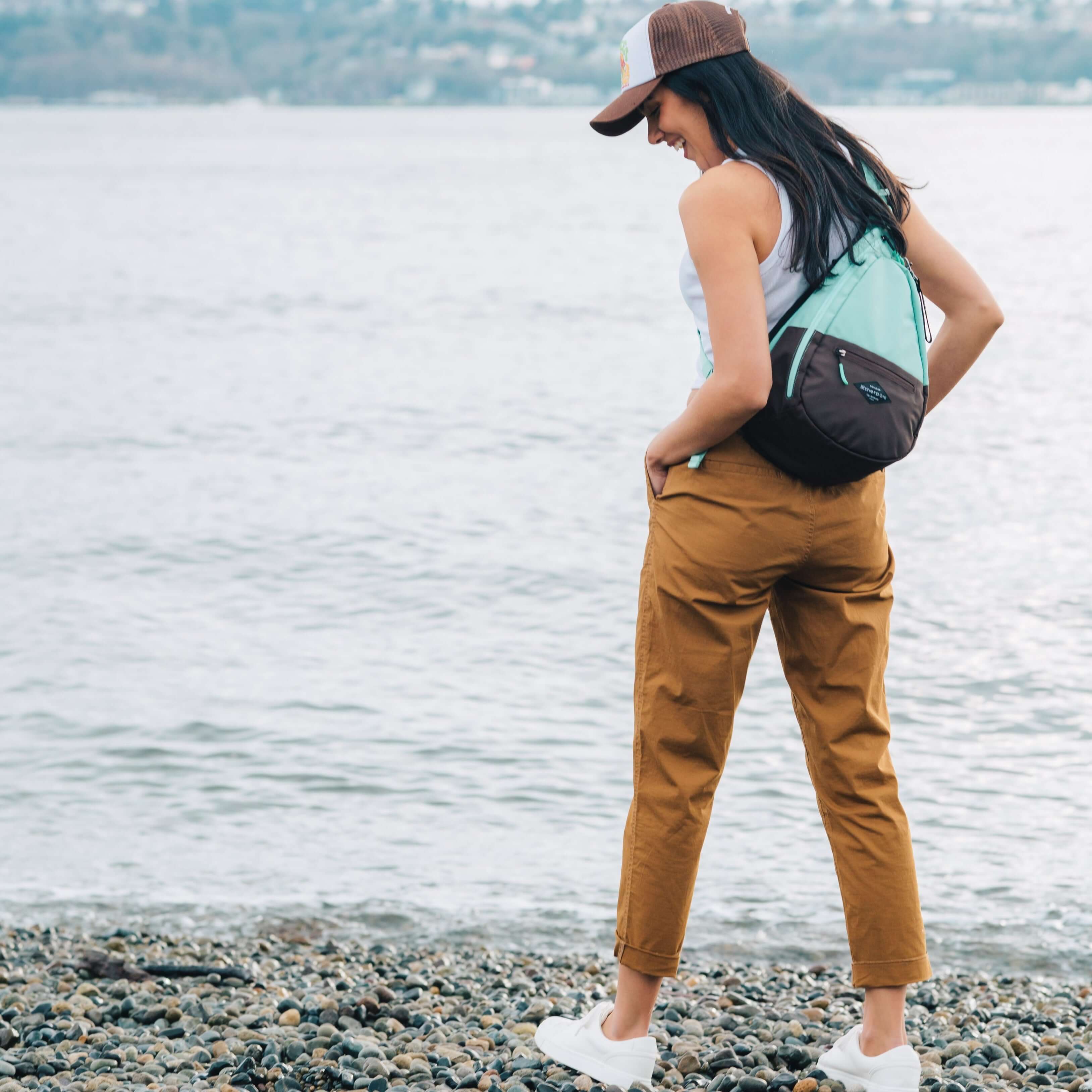 A woman stands outside by a lake, she is facing away from the camera. She is wearing a ball cap, white tank top, brown pants and white shoes. She carries Sherpani bag the Esprit in Seagreen, as a crossbody. 