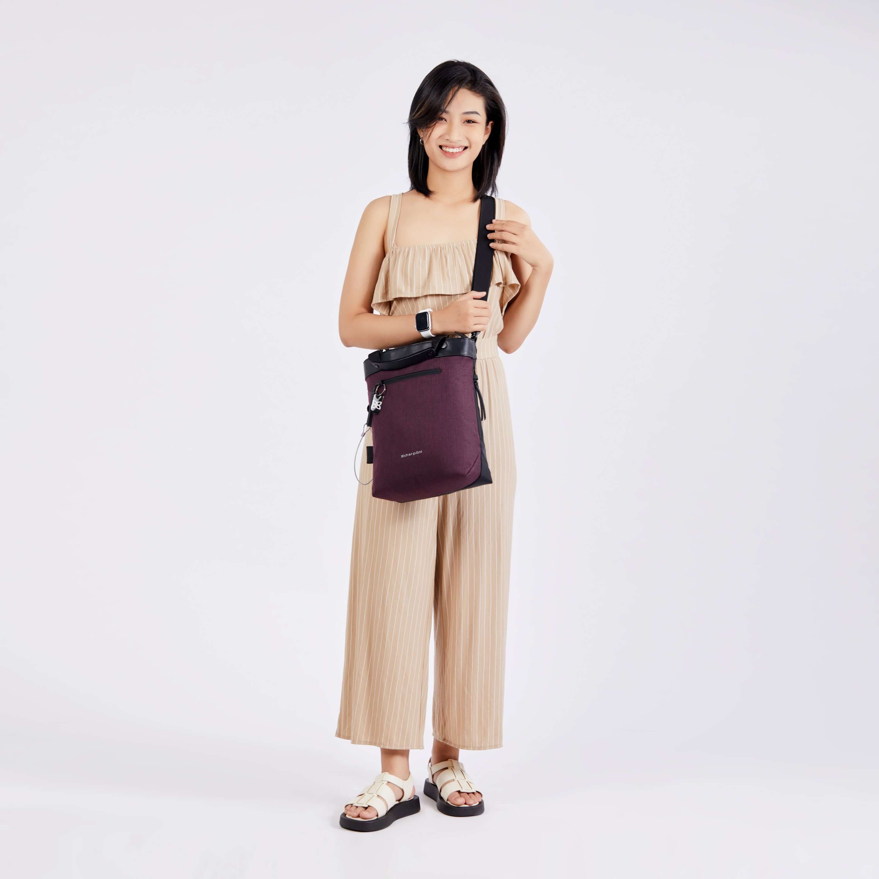 Full body view of a dark haired model facing the camera and smiling. She is wearing a tan pinstriped jumpsuit, white sandals and Sherpani&#39;s Anti-Theft bag, the Geo AT in Merlot, as a crossbody.
